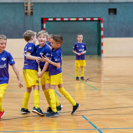 Mehrere junge Jungen in blauen und gelben Trikots feiern auf einem Basketballplatz. Ein Junge steht im Hintergrund in der Nähe des Tores.