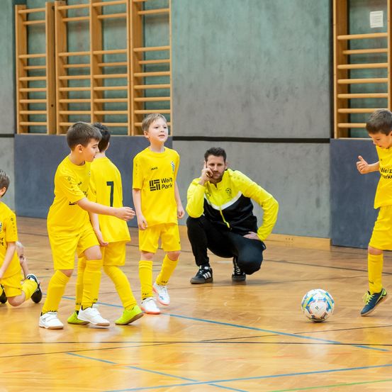 Ein Fußballtrainer weist junge Spieler in gelben Trikots an. Die Kinder beobachten ihn aufmerksam. Der Trainer kniet und spricht am Telefon. Ein Fußball liegt auf dem Boden.