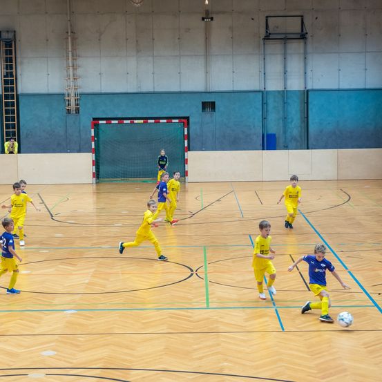 Auf einem Hallenfußballplatz spielen junge Jungen in gelben und blauen Trikots Fußball. Ein Junge in Blau jagt dem Ball hinterher, während andere um ihn herumlaufen. Im Hintergrund steht ein Tornetz.