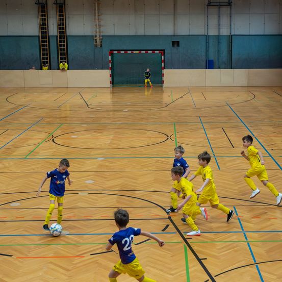 Eine Gruppe junger Jungen in gelben und blauen Trikots spielt Fußball auf einem Hallenplatz, ein Spieler in Blau führt den Ball.