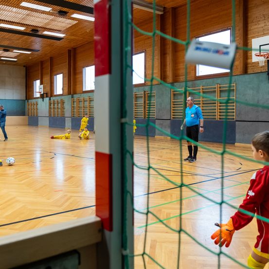 Hallenfußballspiel mit einem jungen Torwart in Rot, Kindern in Gelb, die spielen, und einem Mann in Blau, der als Schiedsrichter fungiert. Holzwände und ein Basketballkorb im Hintergrund.