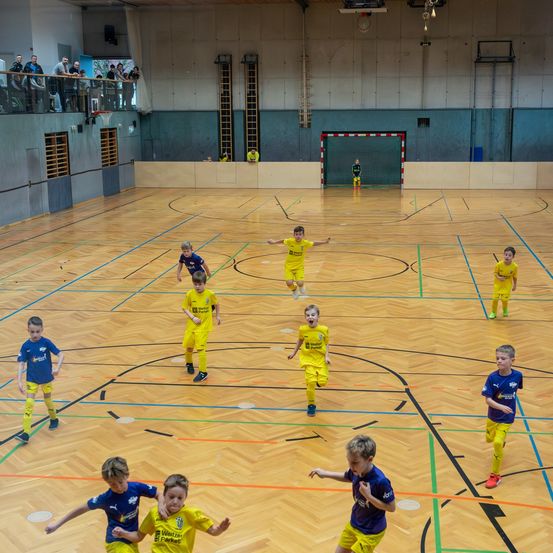 Eine Gruppe junger Kinder spielt Fußball auf einem Hallenplatz. Sie tragen Uniformen. Einige jagen dem Ball hinterher, während andere still stehen. Der Platz hat einen Holzboden mit Markierungen. In der Ferne steht ein Torpfosten.