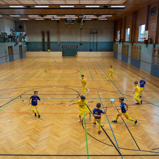 Mehrere Kinder spielen Fußball in einer Halle. Die Kinder tragen Uniformen und Turnschuhe. Der Platz hat einen Holzboden und einen Basketballkorb. An der Wand befinden sich zwei Basketballkörbe.