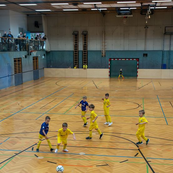 Eine Gruppe junger Jungen in gelben Trikots spielt Fußball auf einem Hallenplatz. Sie rennen und schießen einen Ball. Eine Person steht vor einem Tor. Der Platz hat blaue und orange Markierungen.