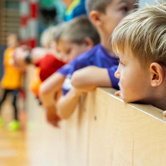 Eine Gruppe von Kindern beobachtet ein Sportereignis hinter einer Holzbarriere. Der Junge vorne schaut zur Seite.