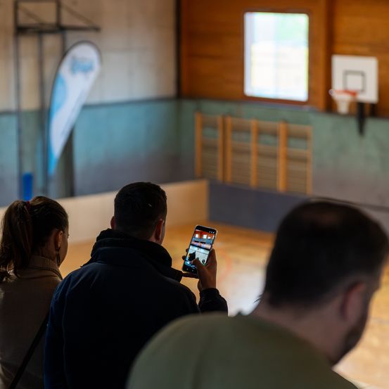 Drei Personen befinden sich in einer Turnhalle, wobei ein Mann ein Handy hält. Der Raum hat Holzwände, einen Basketballkorb und eine Fahne auf der linken Seite.