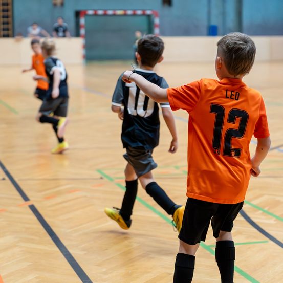 Kinder spielen Hallenfußball; ein Junge in einem orangefarbenen Trikot mit der Nummer 12 zeigt nach vorne.