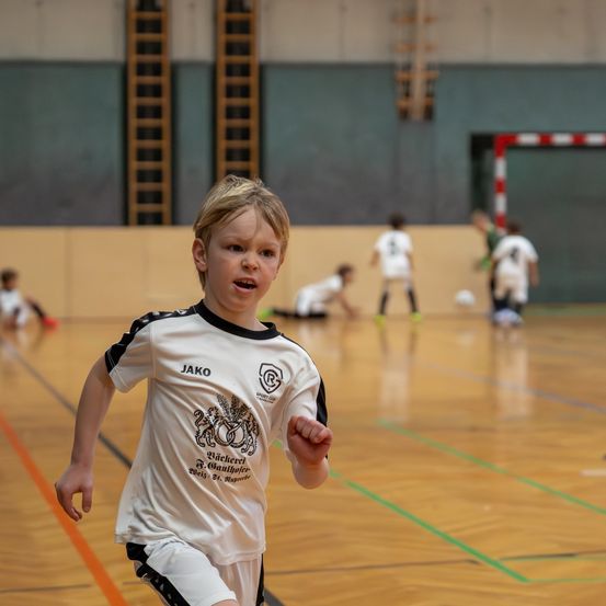 Ein junger Junge läuft auf einem Fußballplatz. Er trägt ein Trikot mit dem Wort Jako. Hinter ihm sind andere Spieler auf dem Platz.