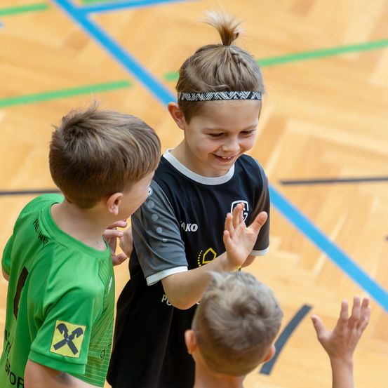 Drei Kinder, zwei Jungen und ein Mädchen, stehen in einer Turnhalle. Das Mädchen lächelt und ein Junge hebt seine Hände.