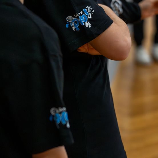Eine Gruppe von Menschen in schwarzen T-Shirts mit einem Club-Logo auf den Ärmeln steht auf einem Holzboden, wahrscheinlich in einer Turnhalle.