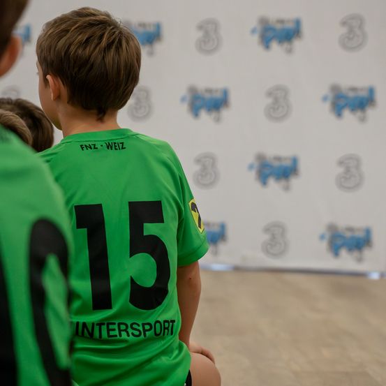 Ein Junge in einem grünen Trikot mit der Nummer 15 sitzt auf dem Boden und blickt nach vorne. Hinter ihm zeigt eine Wand Logos. Ein weiterer Junge in einem ähnlichen Trikot steht teilweise sichtbar.