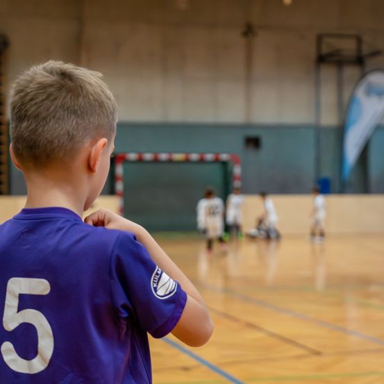 Ein Junge mit der Nummer 5 auf seinem lila Shirt beobachtet ein Spiel in einer Turnhalle mit anderen Spielern und einem Tor.