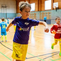 Mehrere junge Jungen spielen Fußball in einer Halle. Ein Junge in einem lila Shirt läuft, während andere hinter ihm stehen oder gehen.
