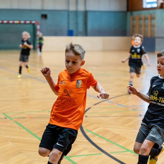 Kinder spielen Hallenfußball, einer in Orange mit 'KIMASOL' auf dem Trikot, der andere in Schwarz mit 'JAKO' auf der Hose. Der Platz hat grüne Markierungen.