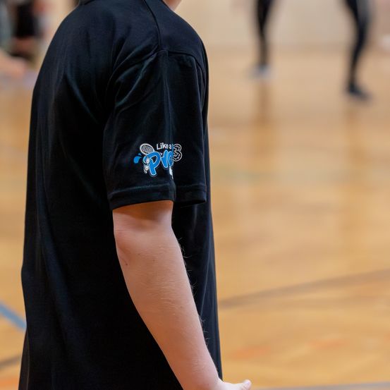 Eine Person in einem schwarzen T-Shirt mit einem blauen Logo auf dem linken Ärmel steht in einer Turnhalle.
