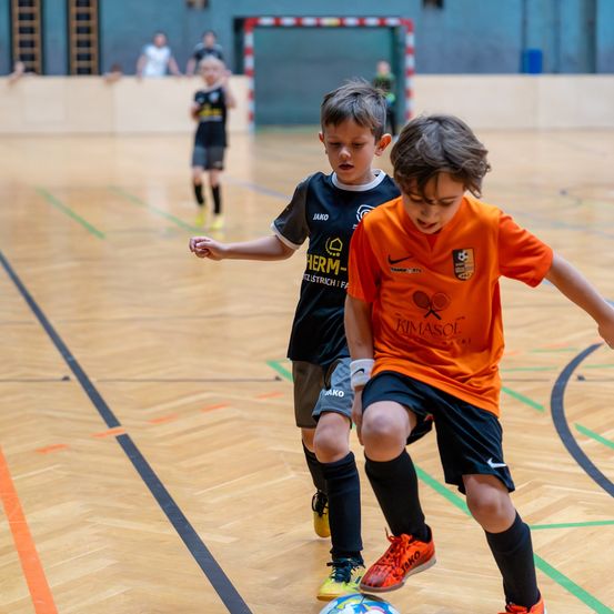 Mehrere junge Jungen spielen Fußball in der Halle. Ein Junge in Orange dribbelt den Ball, während ein anderer Junge in Schwarz verteidigt.