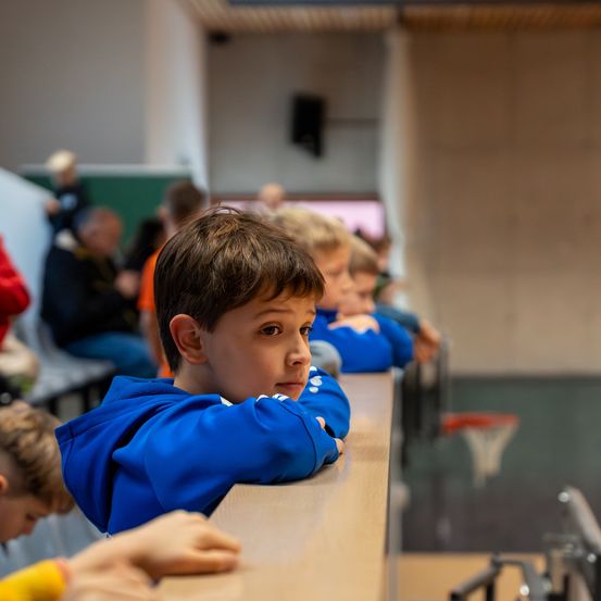 Ein junger Junge in einer blauen Kapuzenjacke lehnt sich an eine Barriere und sieht nachdenklich aus. Hinter ihm sitzen mehrere Kinder, und ein Basketballkorb ist im Hintergrund zu sehen.