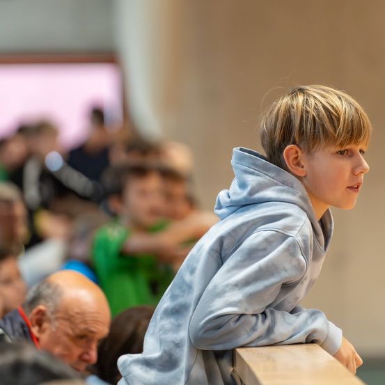 Ein junger Junge in einem hellblauen Hoodie steht in der Nähe einer hölzernen Barriere und schaut in Richtung einer verschwommenen Menschenmenge.