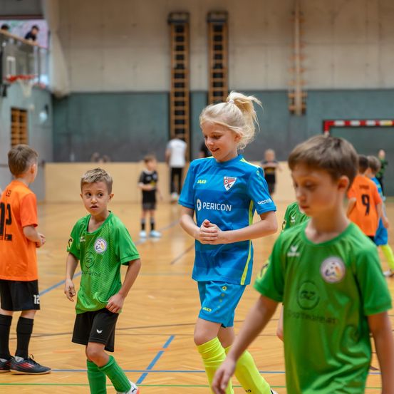 Eine Gruppe von Kindern in Trikots befindet sich auf einem Indoor-Basketballplatz. Ein Mädchen in einem blauen Trikot steht zwischen zwei Jungen in grünen Trikots. Die Kinder spielen wahrscheinlich ein Spiel.