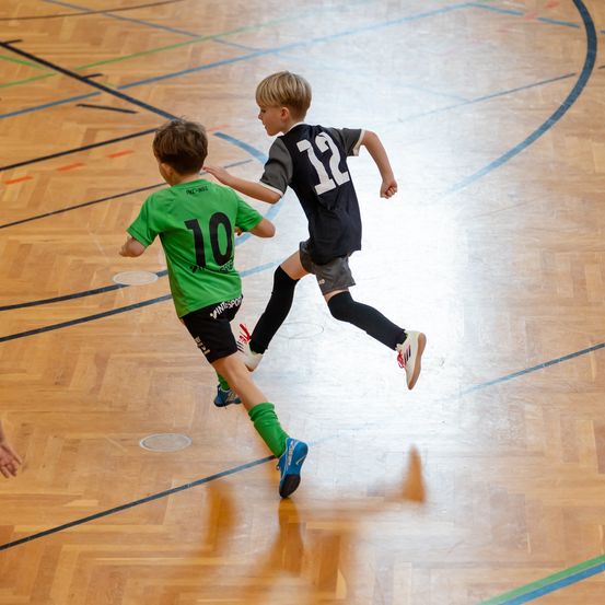 Zwei junge Jungen spielen Hallenfußball, einer trägt ein grünes Trikot mit der Nummer 10 und der andere ein schwarzes mit der Nummer 12. Sie rennen, einer ist leicht voraus.