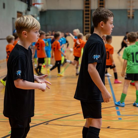 Zwei Jungen in schwarzen Trikots stehen in einer Turnhalle und schauen sich gegenseitig an, während mehrere Kinder im Hintergrund Fußball spielen. Ein Kind trägt ein grünes Trikot.