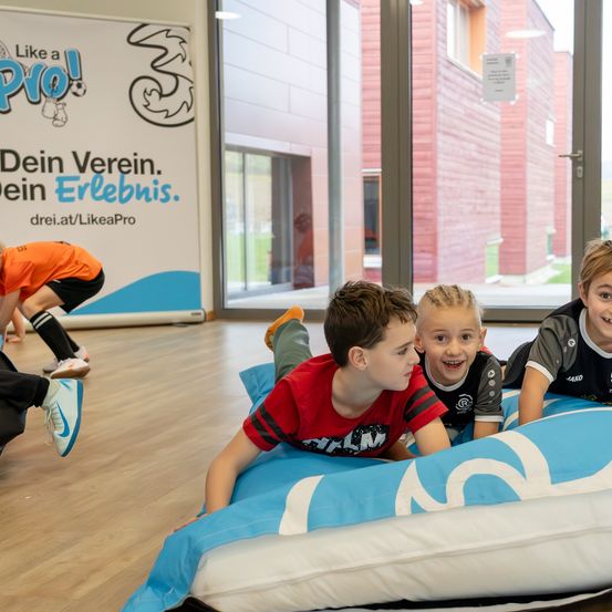 Drei junge Jungen liegen auf einem blau-weißen aufblasbaren Bett. Sie lächeln und schauen nach vorne. Dahinter befindet sich ein großer Banner mit Text und Logo. Eine Person beugt sich nach unten. Ein Gebäude mit Glastüren steht dahinter.