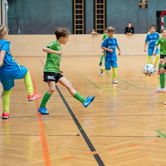 Mehrere Kinder spielen auf einem Hallenplatz Fußball und tragen grüne und blaue Trikots. Ein Kind tritt den Ball, während andere ihm nachlaufen. Zuschauer stehen hinter der Wand.