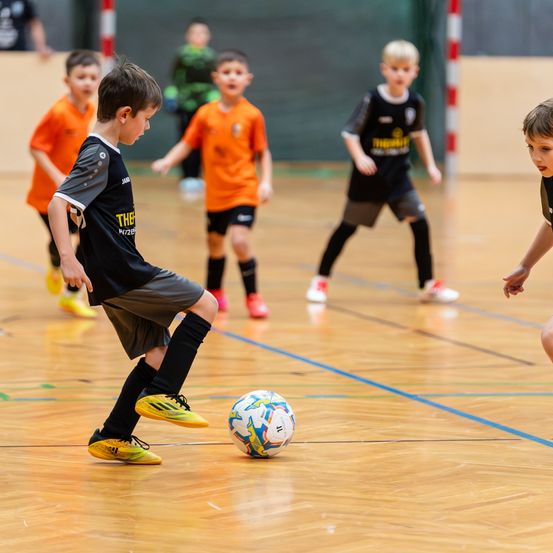 Mehrere junge Jungen spielen Fußball auf einem Hallenplatz. Ein Junge in einem schwarzen Trikot kickt einen Ball, während andere hinter ihm herlaufen. Der Platz hat blaue Linien und eine grüne Wand im Hintergrund.