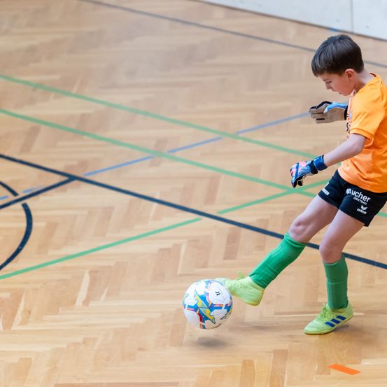 Ein junger Junge in einem orangefarbenen Trikot tritt mit seinem Fuß einen Fußball auf einem Spielfeld.