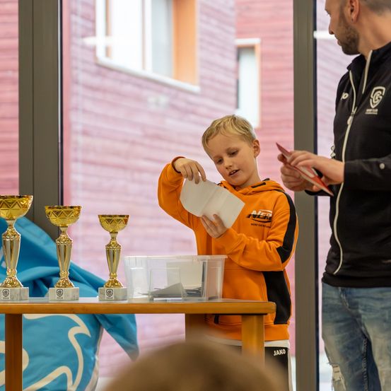 Ein junger Junge in einem orangefarbenen Sweatshirt hält ein Stück Papier in der Hand und steht vor einem Tisch mit Pokalen. Ein Mann in einem schwarzen Sweatshirt steht neben ihm.