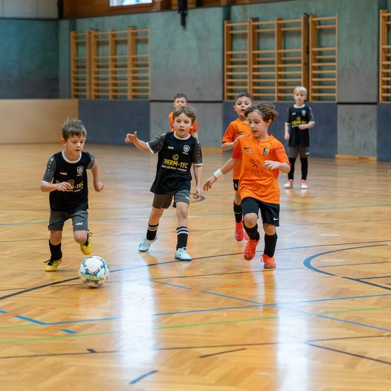 Eine Gruppe junger Jungen in Fußballtrikots spielt eine Partie Fußball in einer Halle. Ein Junge tritt den Ball, während andere ihm folgen.