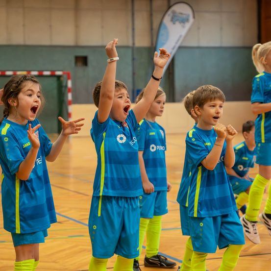 Eine Gruppe von Kindern in blauen und gelben Trikots steht auf einem Basketballplatz und jubelt vor Freude. Hinter ihnen sind eine Flagge und ein Banner angebracht, und ein Torpfosten ist sichtbar. Die Kinder befinden sich in verschiedenen Positionen, wobei einige ihre Hände heben.
