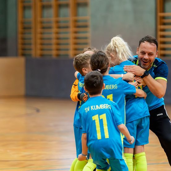 Eine Gruppe junger Athleten in blauen Trikots feiert mit ihrem Trainer in einer Turnhalle. Sie tragen alle Uhren und Turnschuhe.