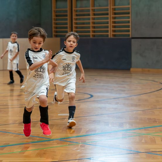 Drei junge Jungen in Sportuniformen laufen in einer Sporthalle. Der Junge vorne hat rote Turnschuhe. Hinter ihnen läuft ein weiterer Junge. Der Boden hat blaue und grüne Markierungen.