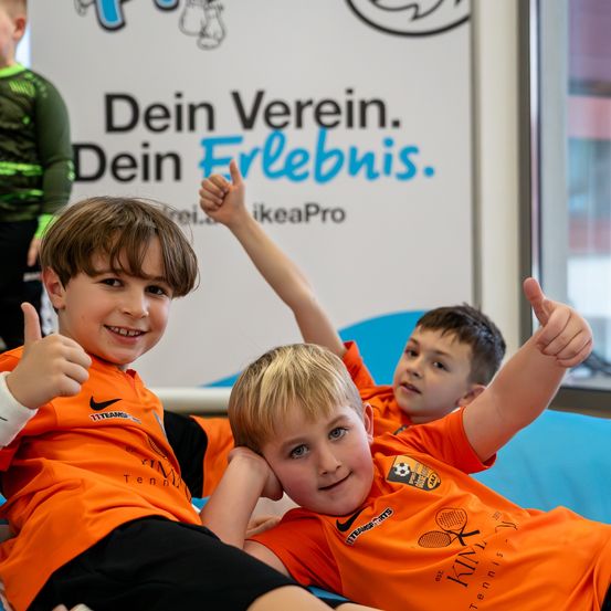 Drei Jungen in orangefarbenen Nike-Sporttrikots sitzen auf einer blauen Oberfläche, lächeln und geben Daumen hoch. Ein Schild im Hintergrund lautet 'Dein Verein. Dein Erlebnis.' Hinter ihnen steht eine Person in einem grünen Outfit.