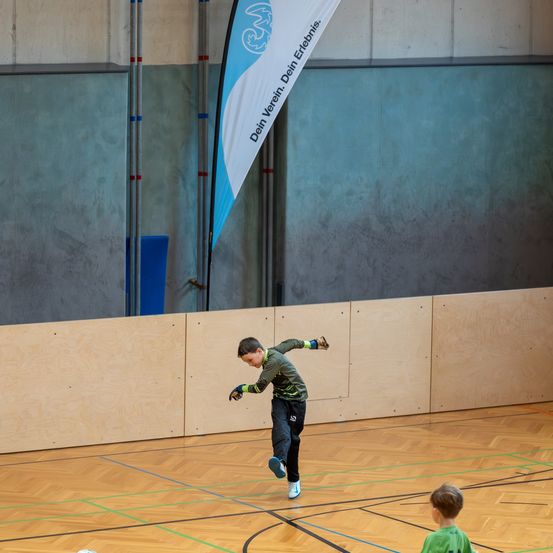 Ein Junge springt auf einem Holzplatz mit einem Fußball, während ein anderes Kind im Hintergrund zuschaut. Ein Banner hängt im Hintergrund.