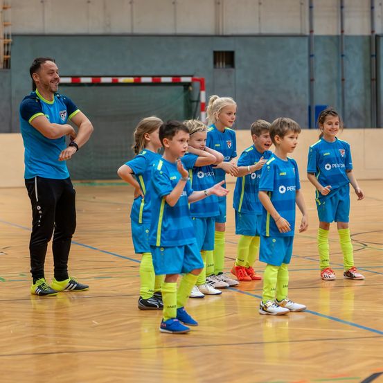 Ein Trainer führt eine Gruppe junger Kinder in blauen und gelben Trikots auf einem Basketballplatz.