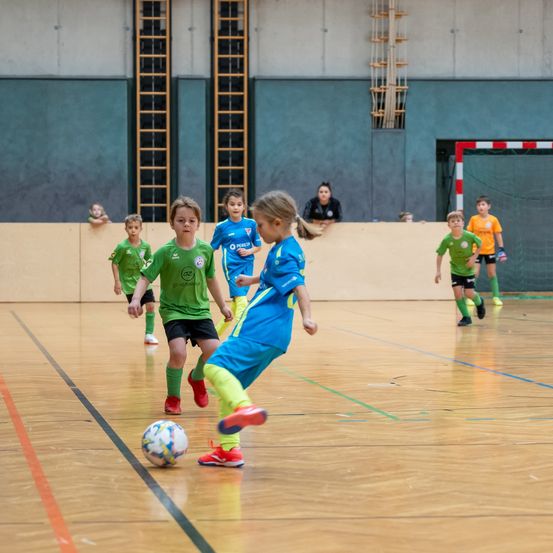 Eine Gruppe von Kindern spielt Fußball in einer Halle. Ein Kind in einem grünen Shirt versucht, den Ball zu treten, während ein anderes Kind in einem blauen Shirt verteidigt. Dahinter beobachtet eine Frau.