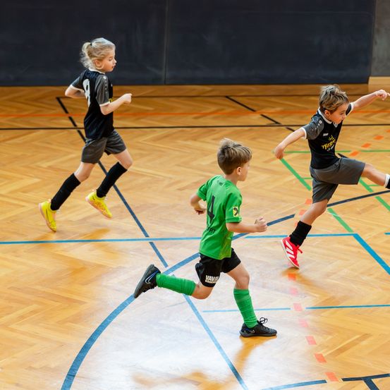 Drei Kinder spielen Fußball auf einem Hallenplatz. Das Kind im grünen Trikot läuft, während die anderen ihm nachjagen.