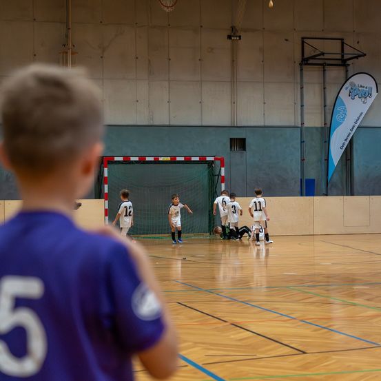 Eine Gruppe junger Jungen spielt Fußball auf einem Indoor-Feld. Ein Junge beobachtet das Spiel.