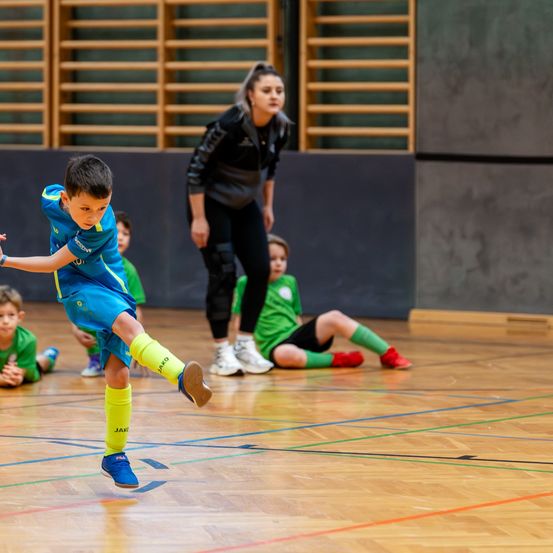 Ein junger Junge spielt drinnen Fußball. Er trägt ein blaues und gelbes Trikot. Eine erwachsene Frau steht hinter ihm.