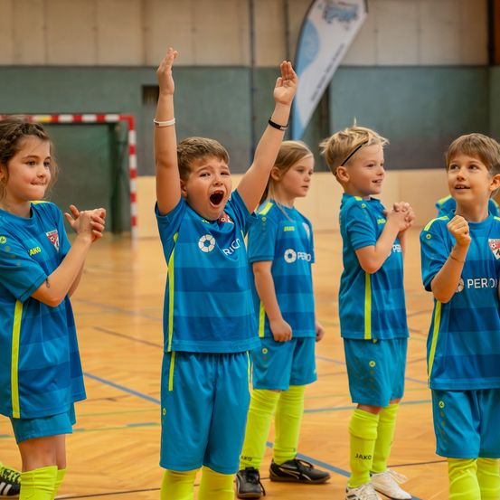 Eine Gruppe von Kindern in blauen und gelben Trikots steht in einer Sporthalle. Sie feiern mit erhobenen Händen.
