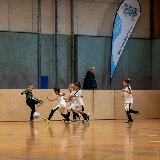 Eine Gruppe junger Jungen spielt Fußball auf einem Indoor-Platz. Ein Junge tritt den Ball, während andere darauf zulaufen. Ein erwachsener Mann steht und beobachtet sie.