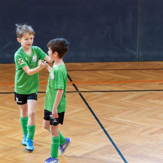 Zwei Jungen in grünen Fußballtrikots schütteln sich auf einem Spielfeld die Hände. Ein Junge hat ein gelbes 'X' auf seinem Trikot.
