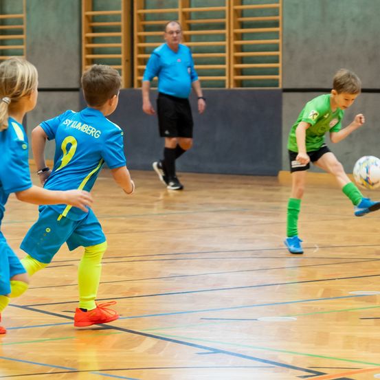 Mehrere Kinder spielen Fußball in einer Turnhalle. Ein Kind in einem grünen Trikot tritt den Ball, während ein anderes Kind darauf zuläuft. Ein Mann in einem blauen Shirt beobachtet sie.