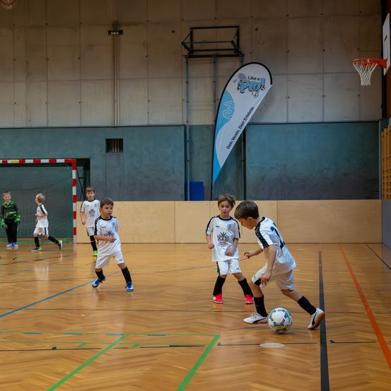 Eine Gruppe junger Jungen spielt Fußball auf einem Hallenplatz. Sie tragen weiße Trikots und Turnschuhe. Ein Spieler ist kurz davor, den Ball zu schießen. Der Platz hat blaue Markierungen.