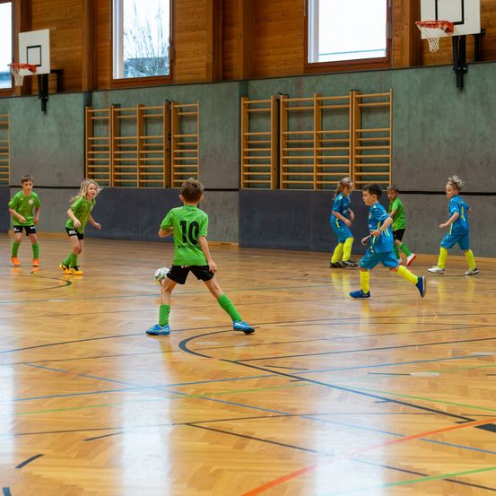 Kinder in Sportuniformen spielen Fußball auf einem Innenfeld. Ein Spieler in einem grünen Trikot mit der Nummer 10 jagt dem Ball hinterher.