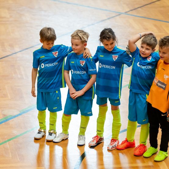 Fünf Kinder in blauen und gelben Sportuniformen stehen zusammen auf einem Spielfeld, zwei tragen Turnschuhe und drei Socken. Sie alle lächeln und schauen in die Kamera.