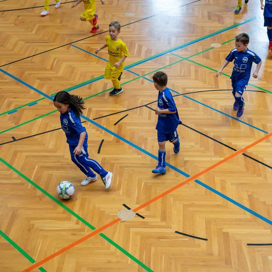 Eine Gruppe junger Kinder, die auf einem Holzboden mit bunten Markierungen drinnen Fußball spielen. Die Kinder tragen blaue und gelbe Trikots.