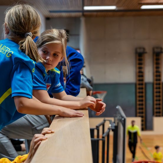 Drei junge Mädchen in blauen Trikots lehnen sich an eine Holzbarriere in einer Turnhalle. Eine schaut nach unten, eine andere schaut zur Seite und eine dritte schaut in Richtung eines Basketballkorbs.